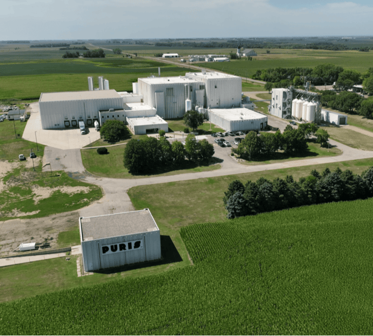 The image shows an aerial view of a large industrial facility with multiple buildings, some of which are labeled 'DURIS'. There are large cylindrical storage tanks, a few vehicles, and the surrounding area has green fields.