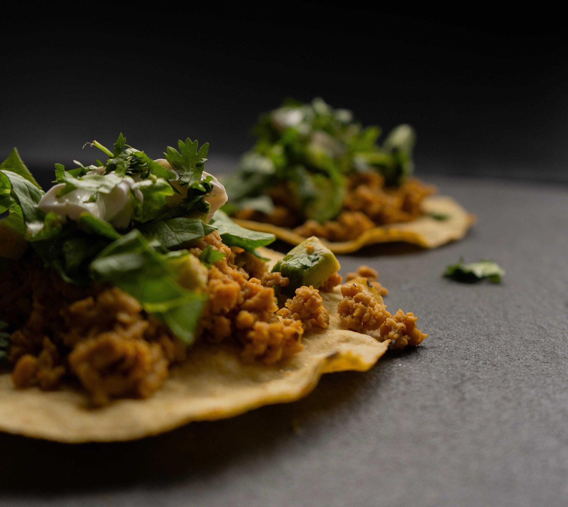 Corn tortilla tostadas topped with seasoned plant-based protein crumbles, avocado, fresh cilantro, and sour cream on a dark surface